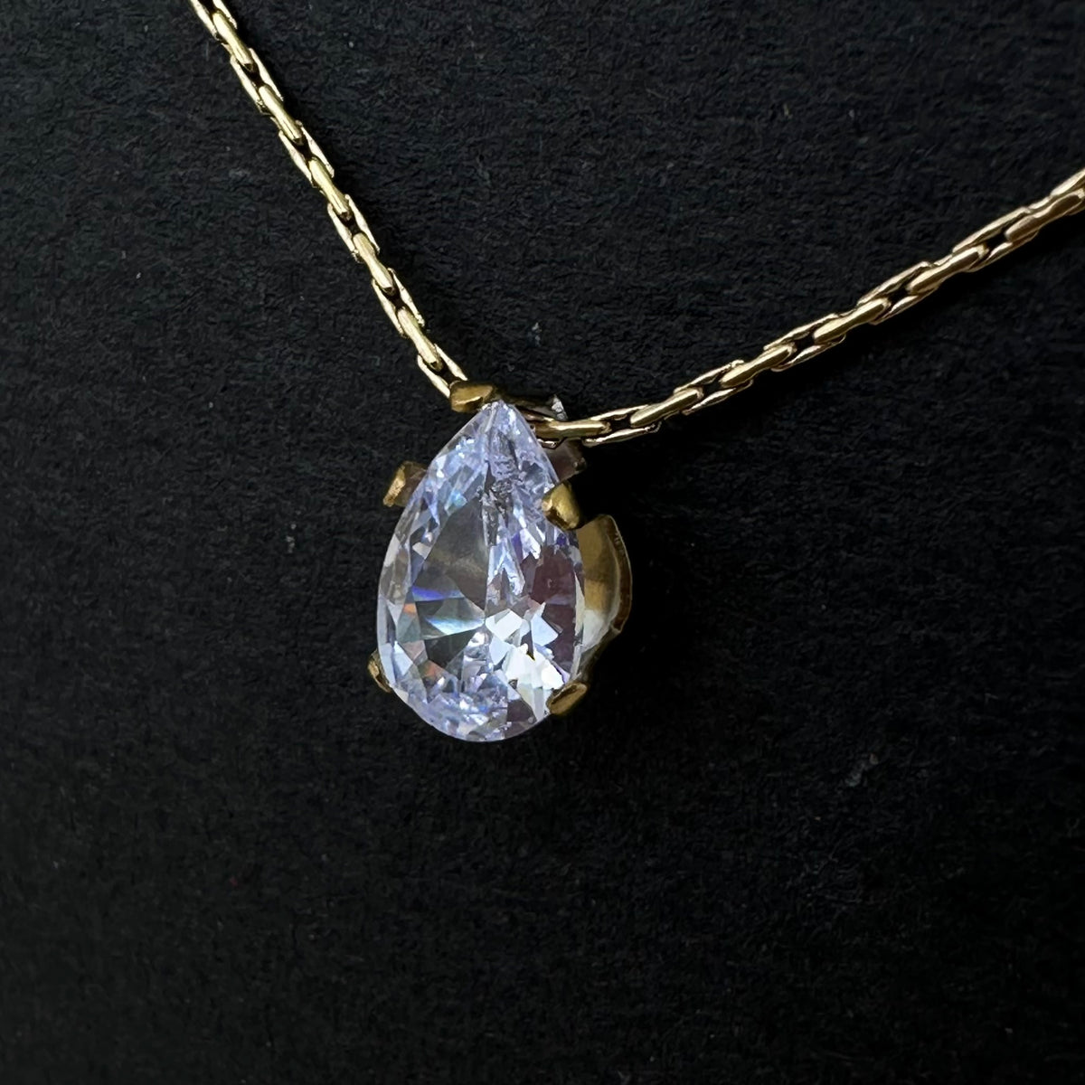 A gold chain necklace with a teardrop-shaped clear gemstone pendant in a gold setting, displayed on a textured black background.