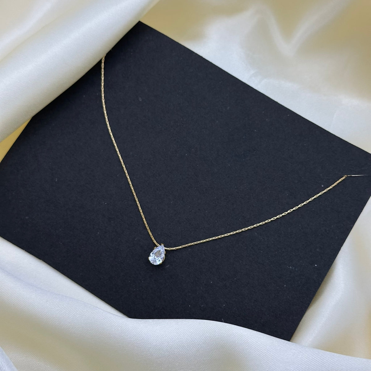 A delicate gold chain necklace with a small teardrop-shaped clear gemstone pendant, displayed on a black square card against a draped cream satin fabric background.