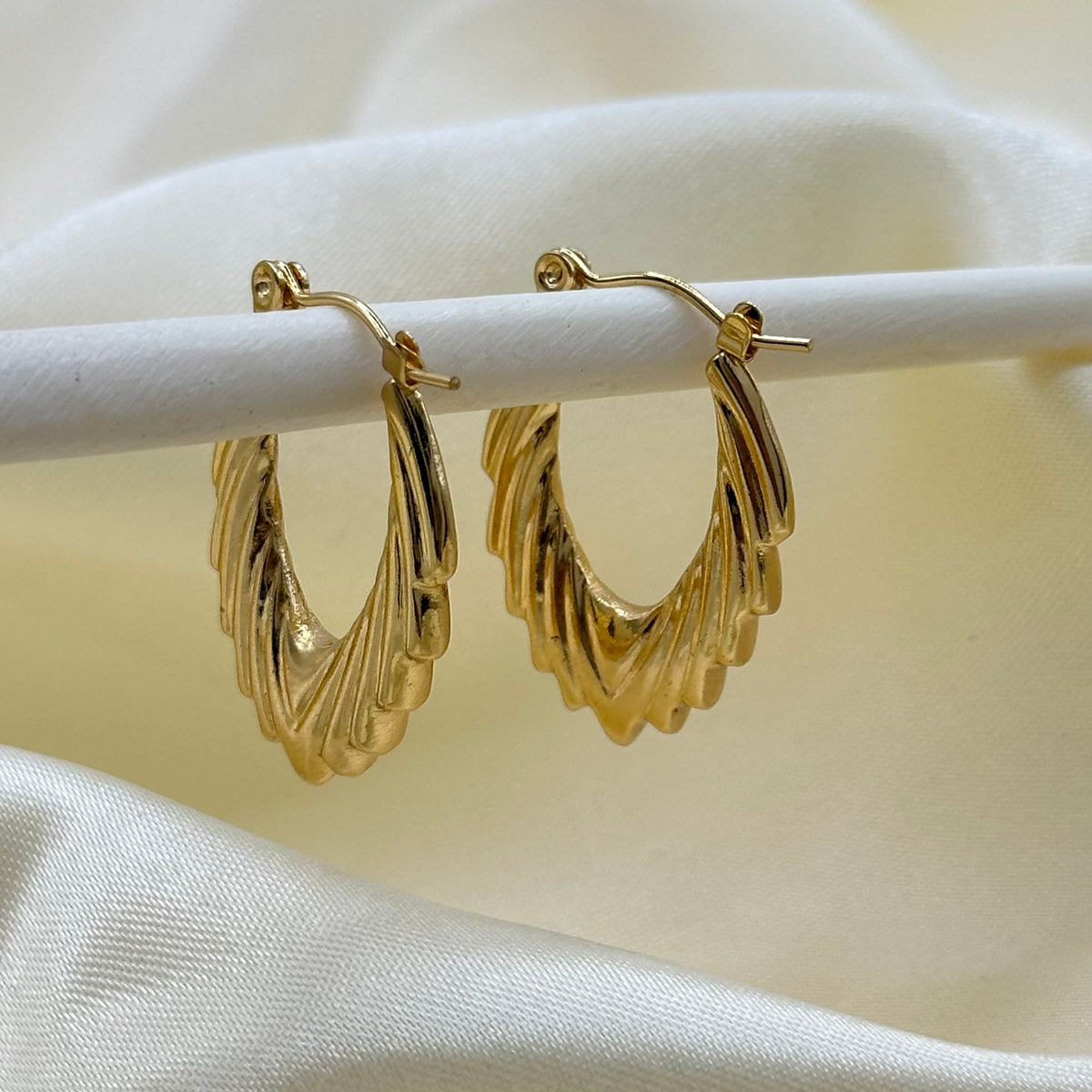 A pair of gold hoop earrings with a textured, layered wavy design, displayed on a white stand against a draped cream satin fabric background.