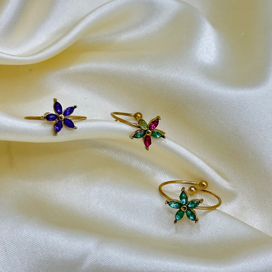 Three gold-tone flower-shaped rings with colorful gemstone accents in purple, green, and multicolour, arranged on draped cream satin fabric.