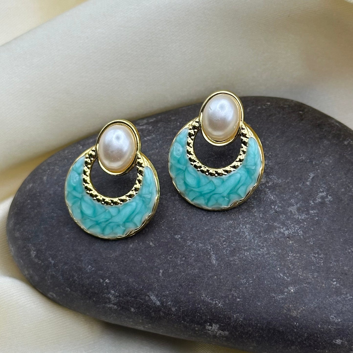 A pair of gold-tone statement earrings with oval white pearls and turquoise-blue marbled enamel discs, displayed on a dark stone against a cream fabric background.