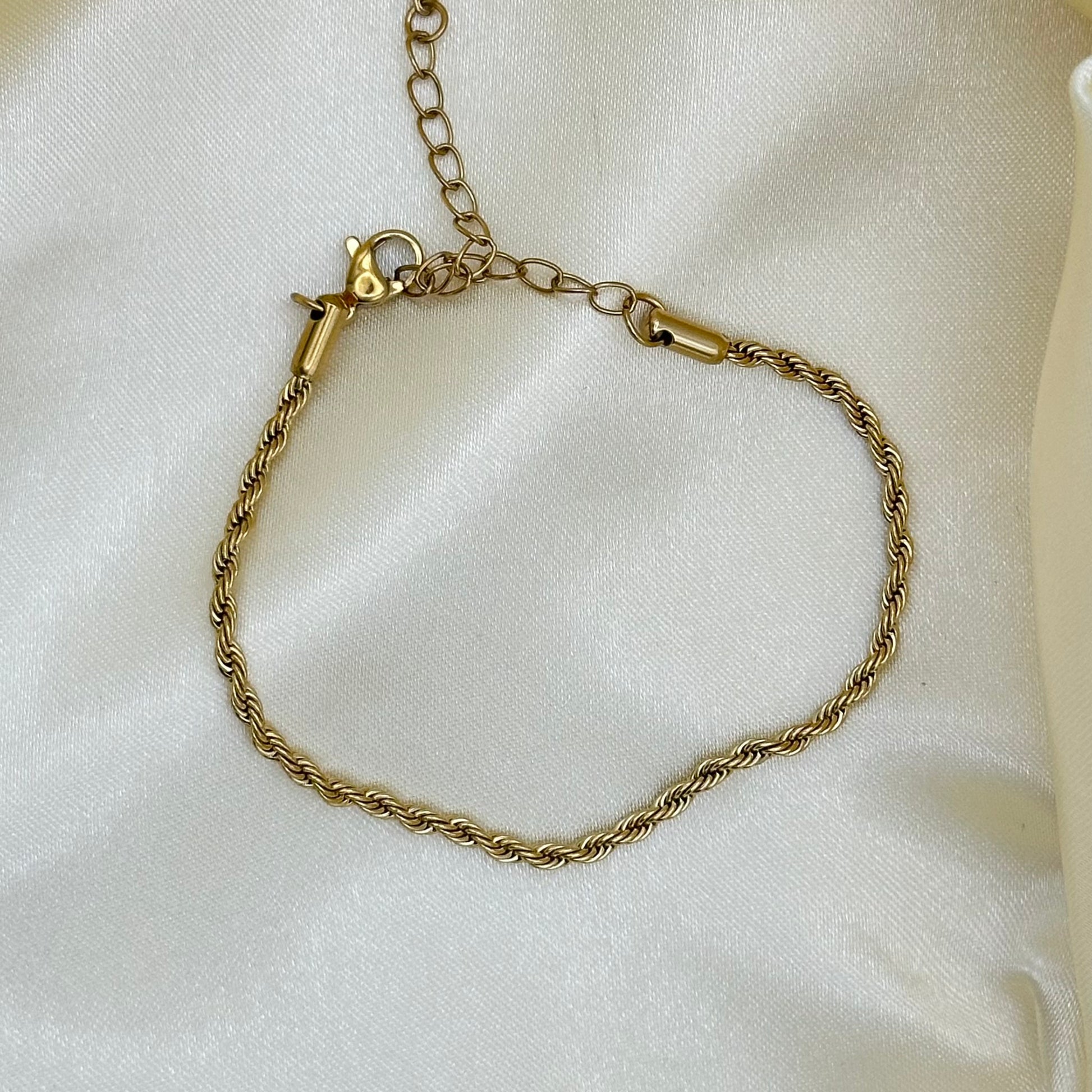 A gold-toned twisted rope chain bracelet with an adjustable lobster clasp and extension chain, displayed on a draped white satin fabric background.