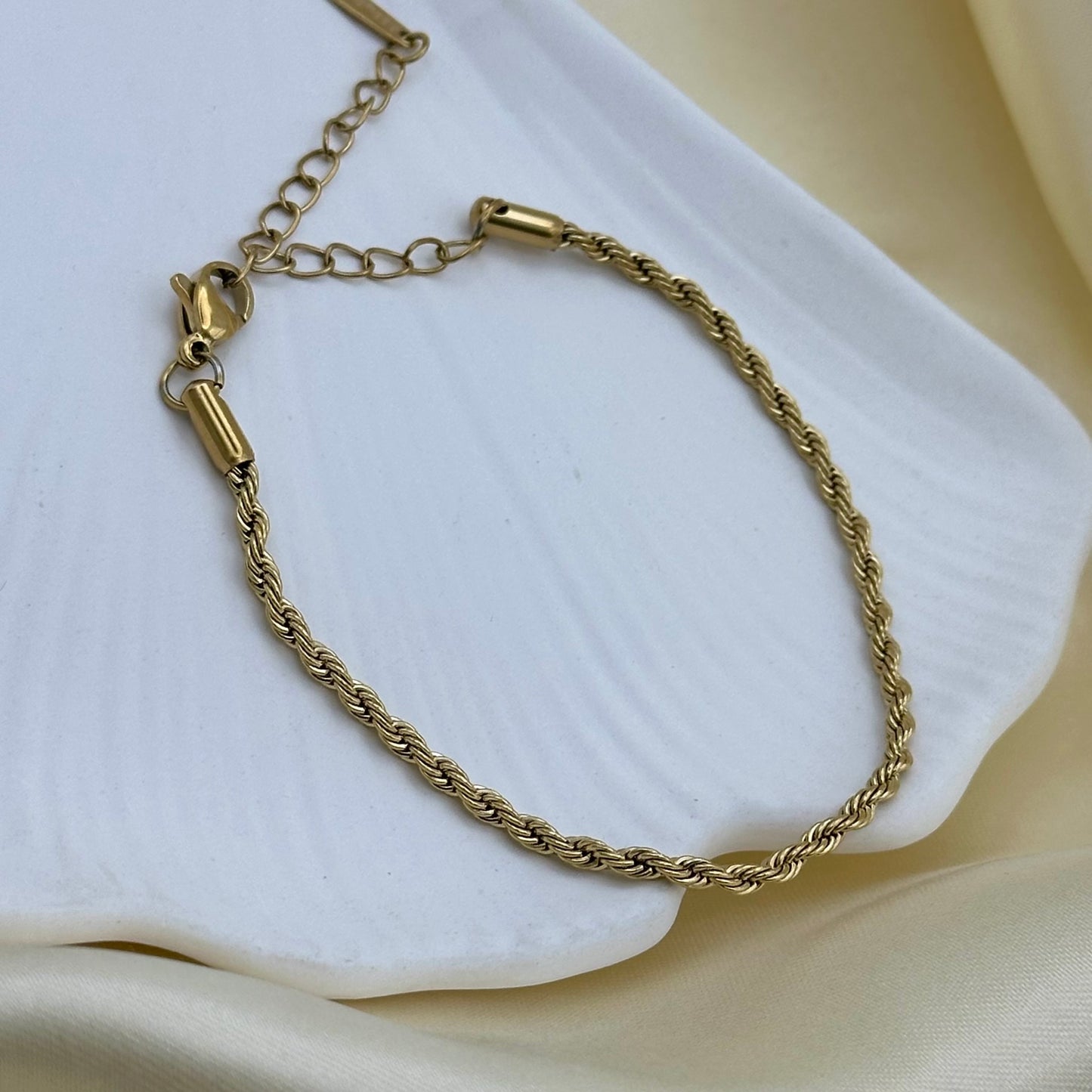 A gold rope chain bracelet with a lobster clasp and extender chain, displayed on a textured white ceramic dish over a soft beige fabric background.