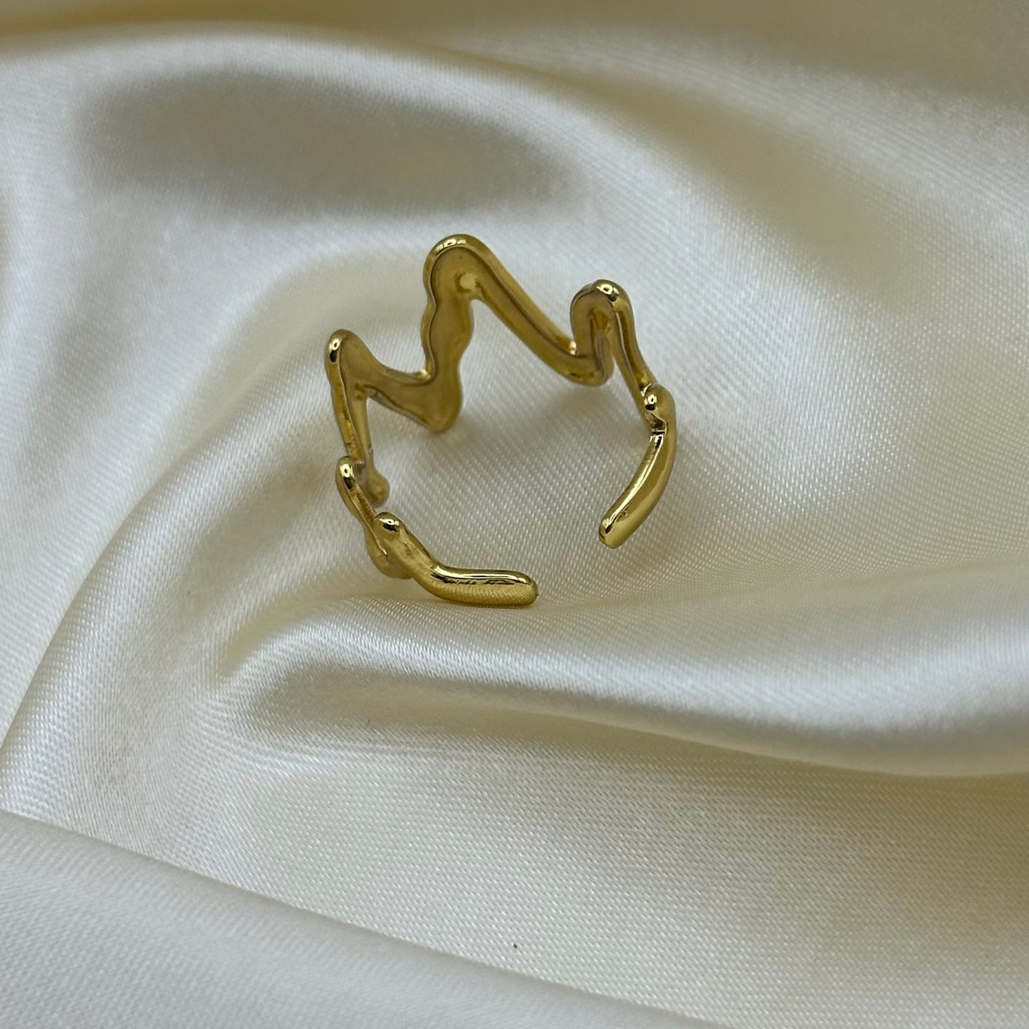 A gold open ring with a wavy, abstract line design, displayed on a draped cream-colored satin fabric background.