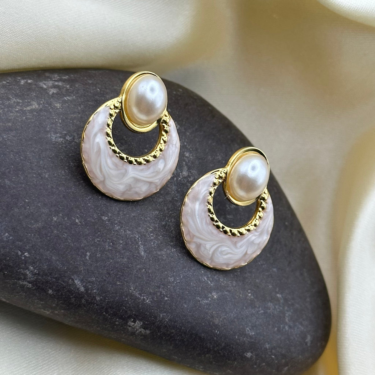 A pair of gold and pearl drop earrings with carved white agate crescent moons, displayed on a dark stone against a cream satin background.