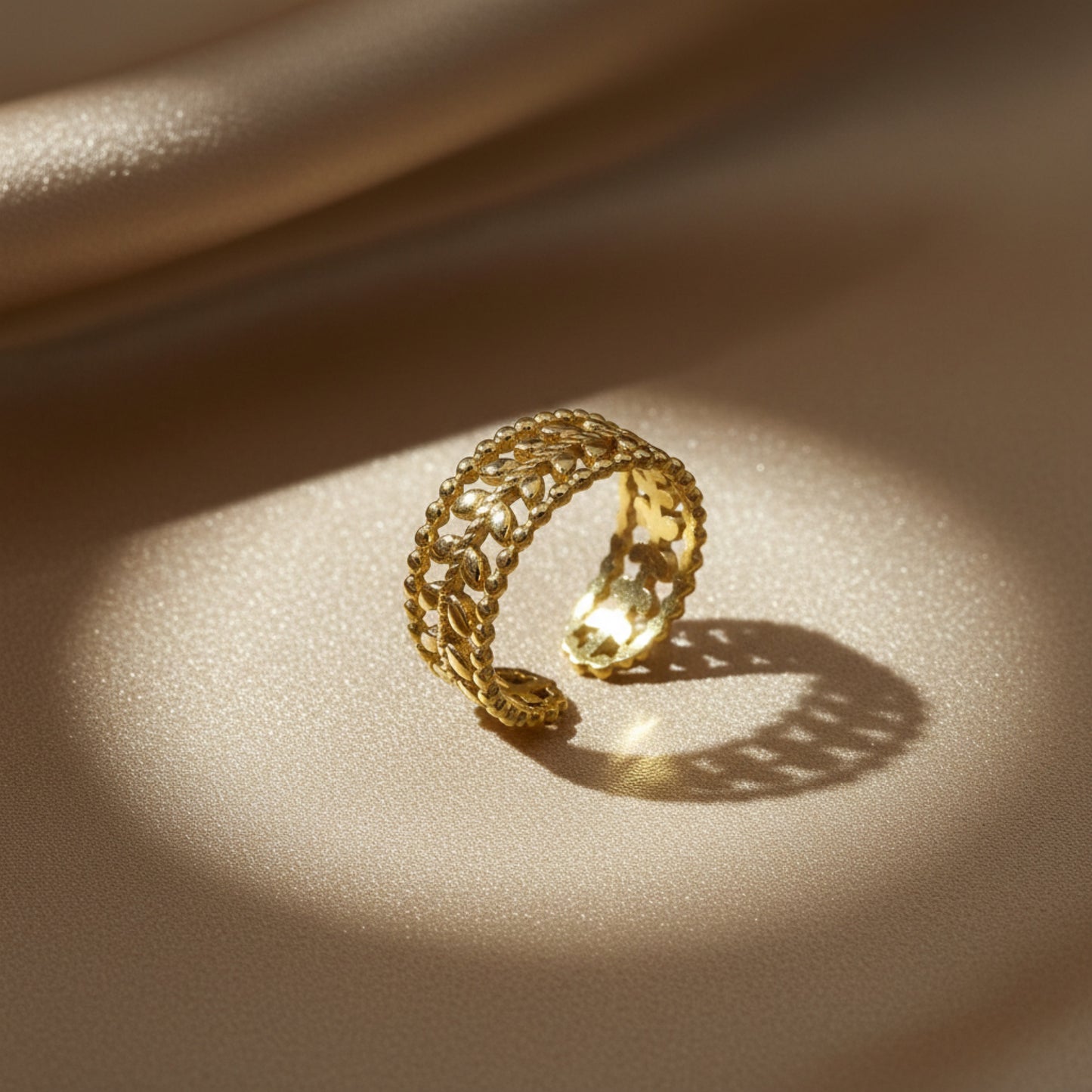 A gold open ring with a textured, woven leaf-like pattern, resting on a shimmering beige surface under dramatic, warm sunlight and shadow.