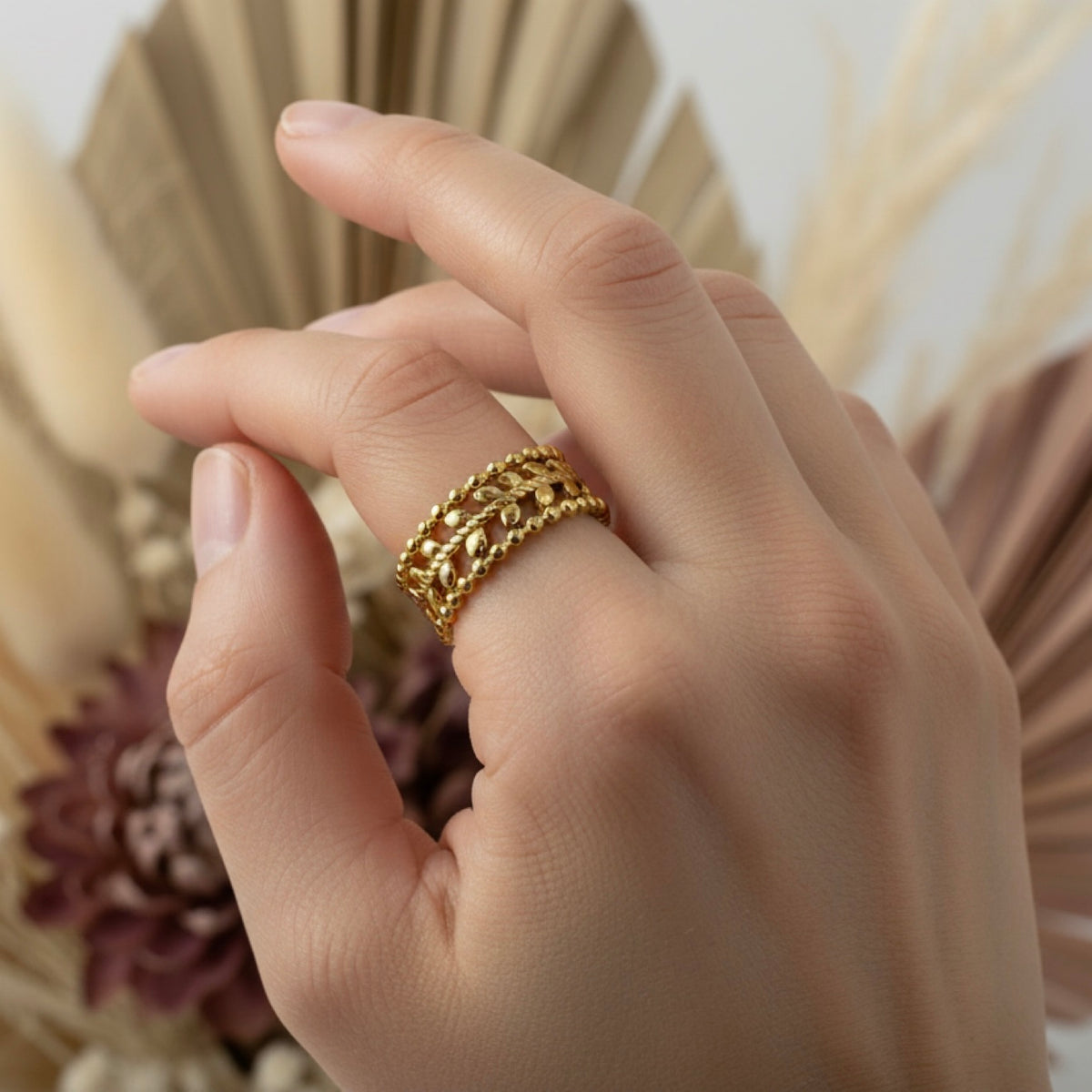 A close-up of a hand wearing a wide, textured gold ring with a woven or braided openwork design, set against a soft blurred background of dried beige and mauve botanical elements.