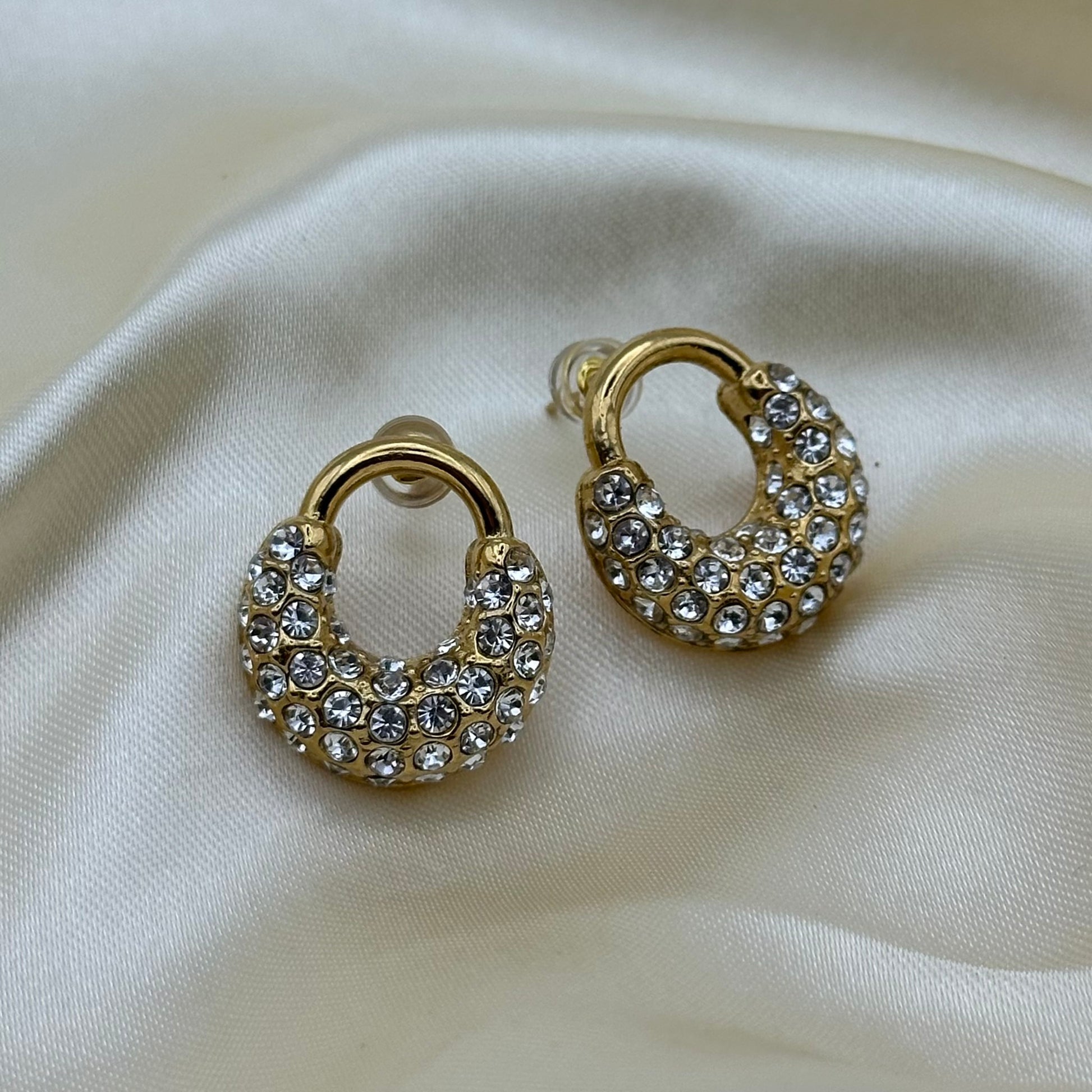 A pair of gold crescent-shaped hoop earrings covered in clear rhinestones, displayed on a draped cream satin fabric.