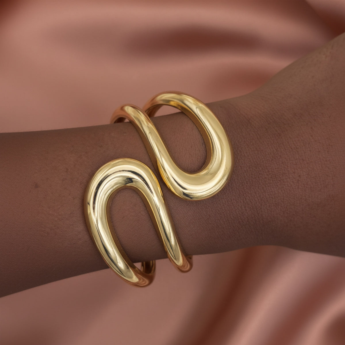A close-up of a polished gold wrap cuff bracelet worn on a person's wrist, set against a soft, draped dusty rose satin background.