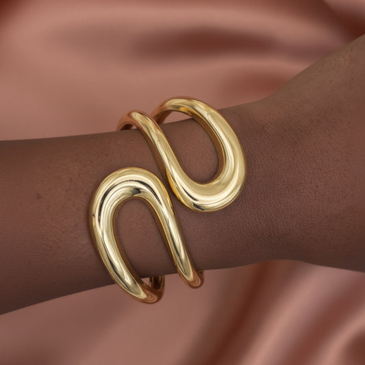 A close-up of a polished gold wrap cuff bracelet worn on a person's wrist, set against a soft, draped dusty rose satin background.