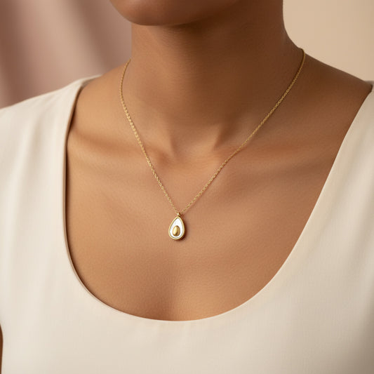 A close-up of a model wearing a delicate gold chain necklace with a teardrop-shaped pendant featuring a white and gold design, paired with a cream-colored scoop-neck top.