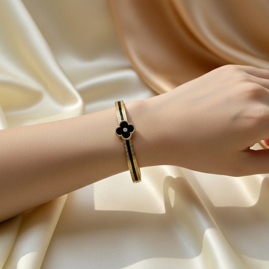 A gold-toned bangle bracelet with a black enamel four-petal flower charm and a small white center, worn on a person's wrist against a draped cream satin background.