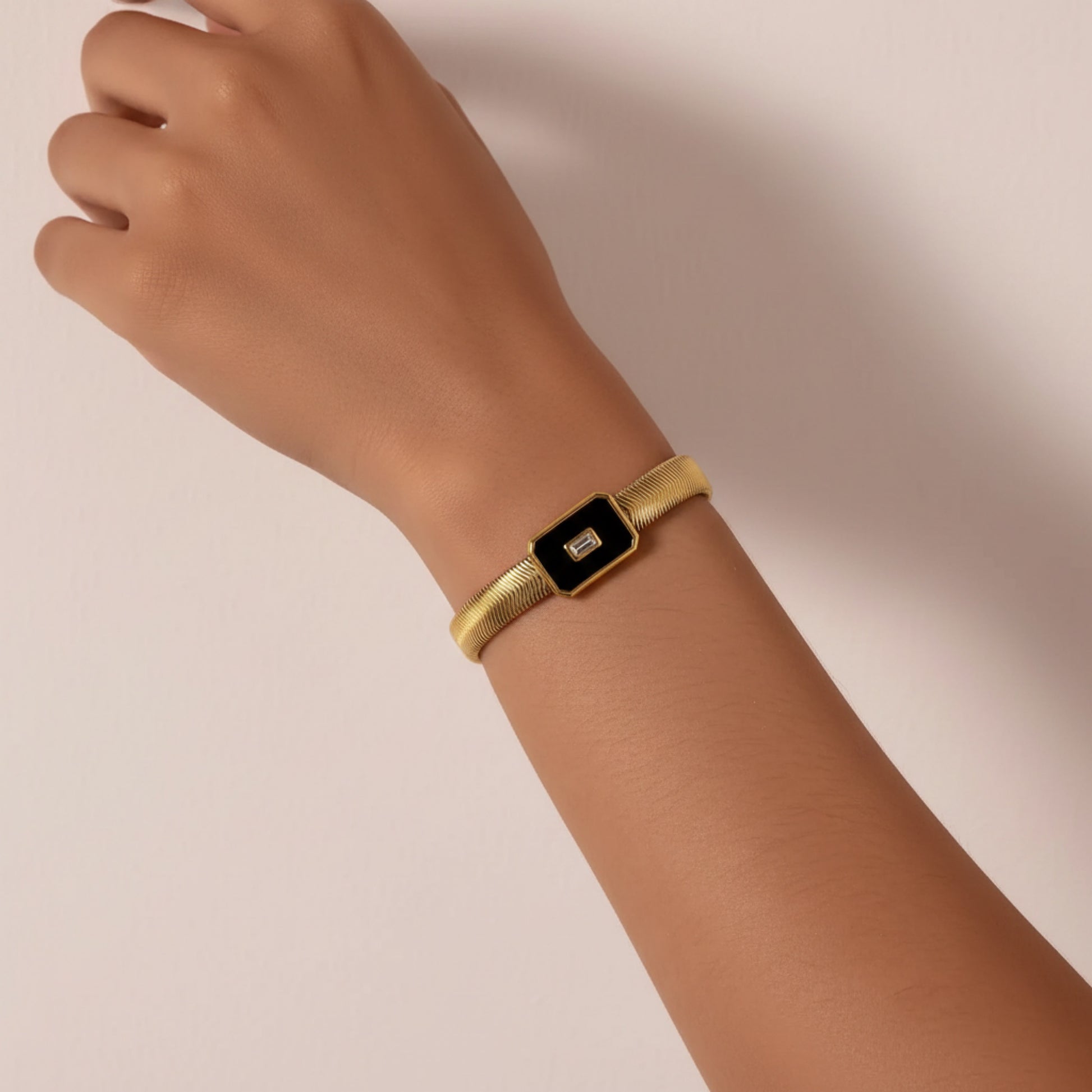 A gold snake chain bracelet with a rectangular black inlay and a small central crystal, shown worn on a person's wrist against a soft beige background.