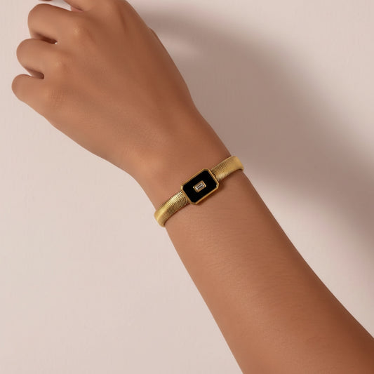 A gold snake chain bracelet with a rectangular black inlay and a small central crystal, shown worn on a person's wrist against a soft beige background.
