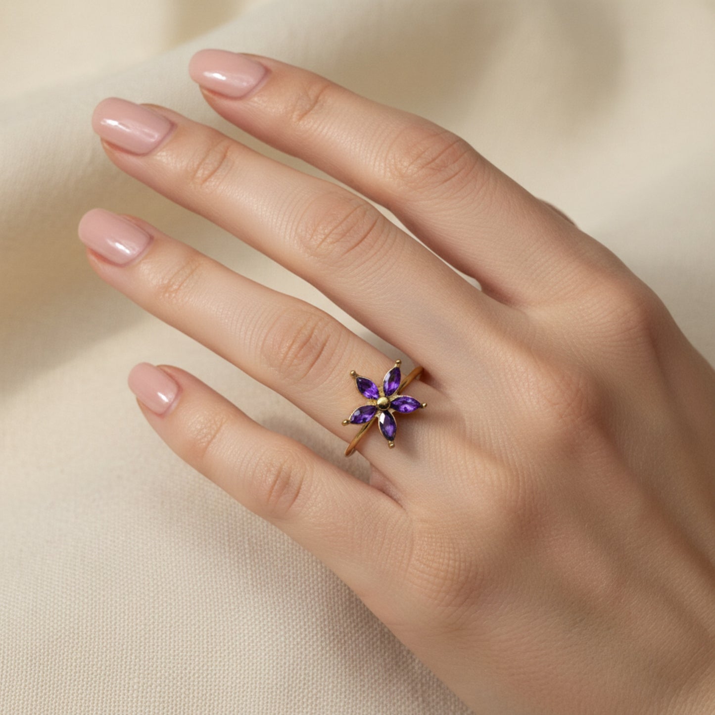 A close-up of a hand wearing a gold ring with a purple crystal flower on the ring finger, against soft cream fabric.