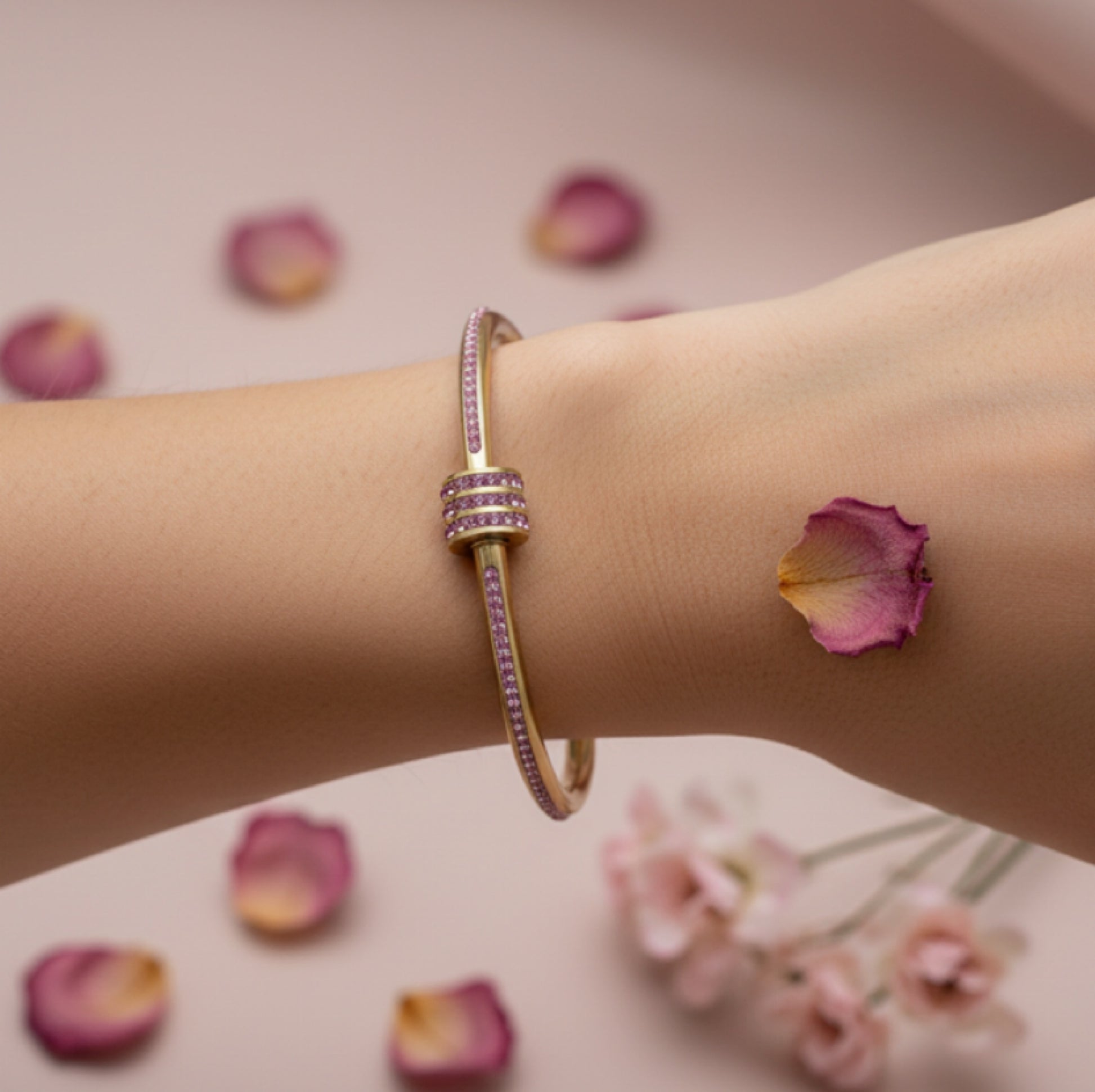 A gold-toned bangle bracelet with pink gemstone accents, shown worn on a person's wrist. The background is a soft pink with scattered rose petals and blurred flowers.