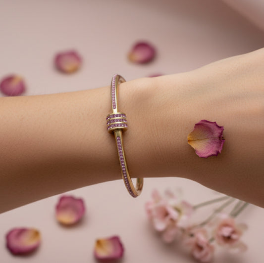 A gold-toned bangle bracelet with pink gemstone accents, shown worn on a person's wrist. The background is a soft pink with scattered rose petals and blurred flowers.