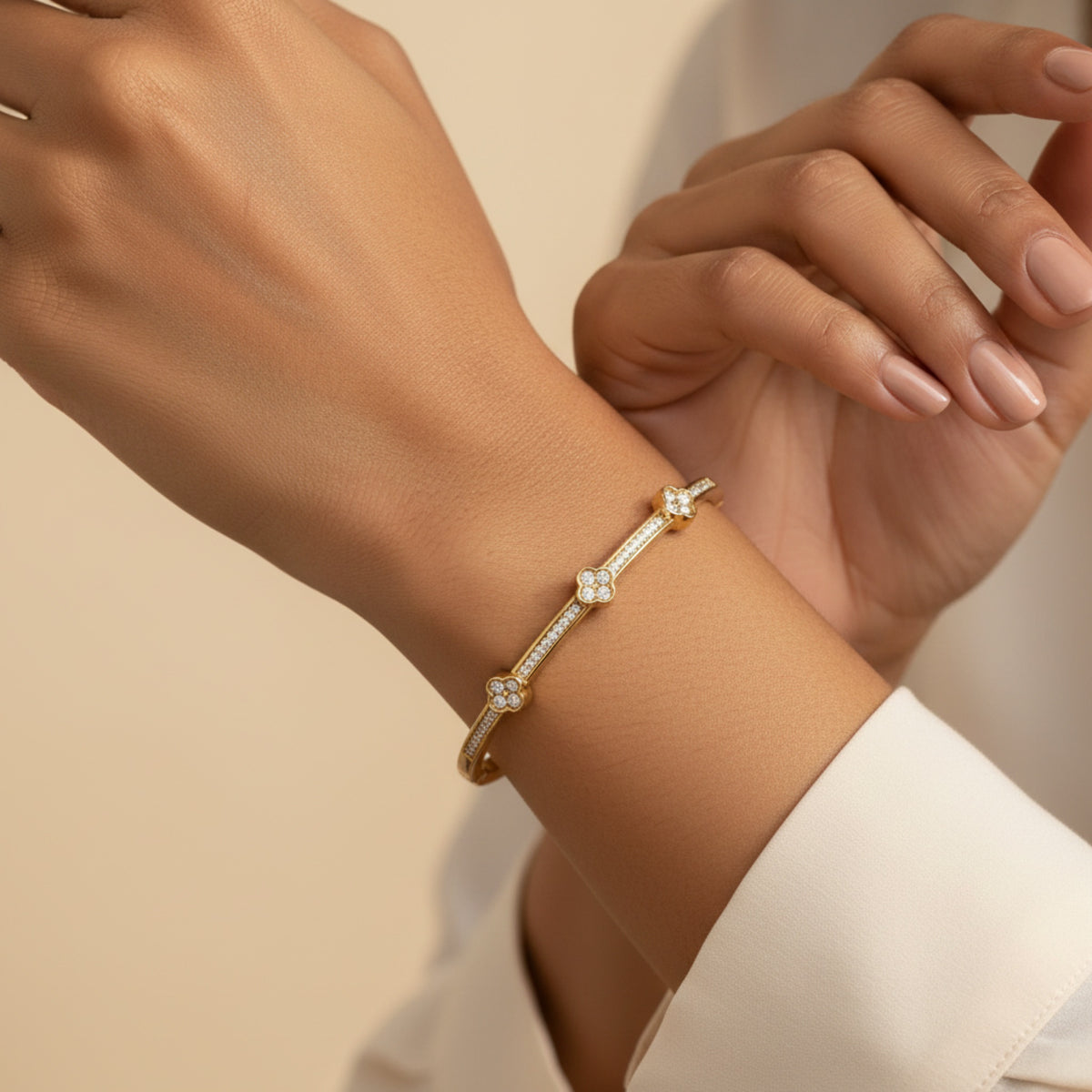 A close-up of a delicate gold bangle bracelet with small clear gemstone accents, worn on a person's wrist. The person is wearing a white collared shirt, and the background is a soft, warm beige.