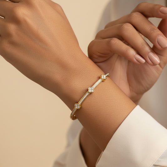 A close-up of a delicate gold bangle bracelet with small clear gemstone accents, worn on a person's wrist. The person is wearing a white collared shirt, and the background is a soft, warm beige.
