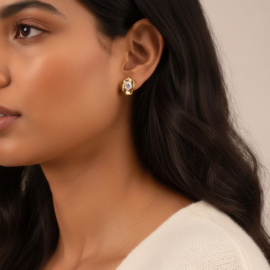 A close-up of a woman wearing a small gold earring with a clear central gemstone. The image shows her ear, hair, and part of her cream-colored top against a soft beige background.