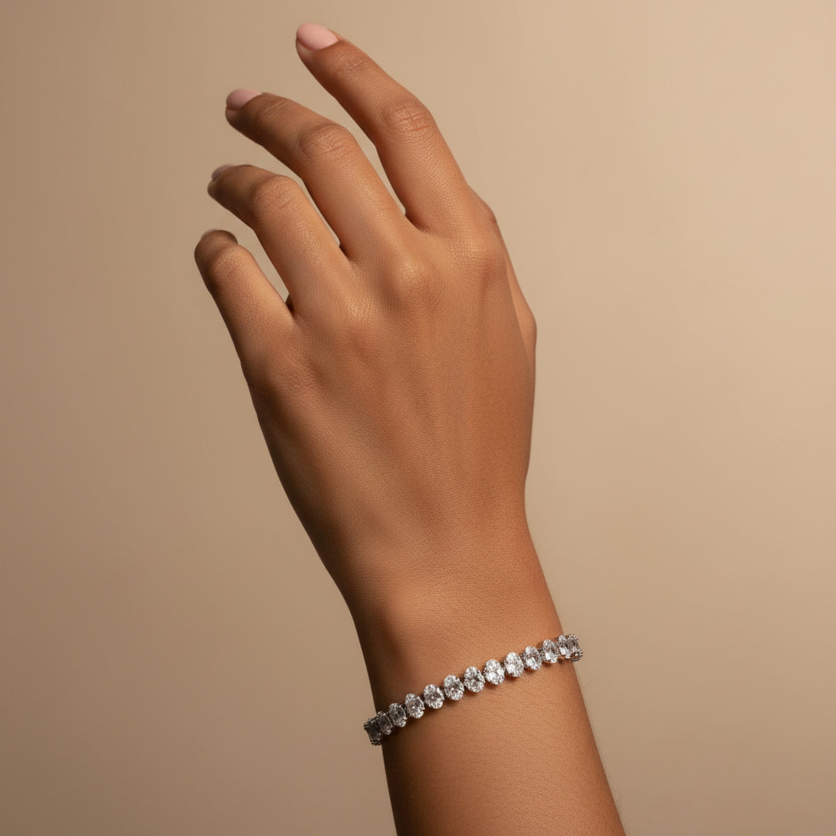 A close-up of a person's hand and wrist wearing a delicate silver-toned bracelet set with clear gemstones, set against a solid warm beige background.