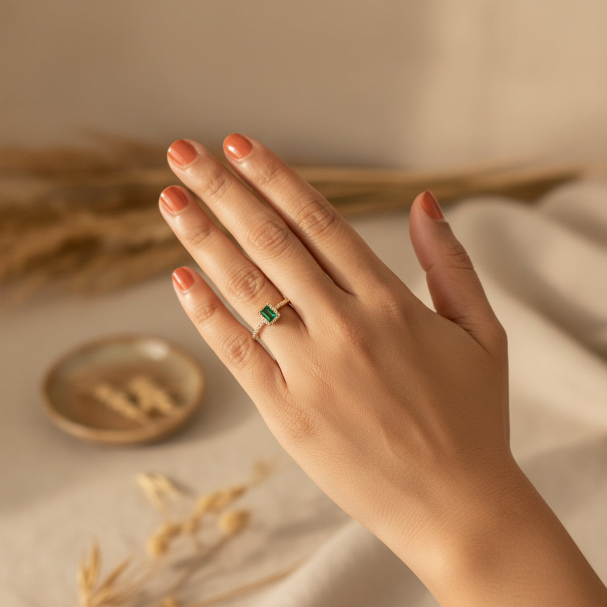 A hand with peach-colored nail polish wearing a delicate gold ring with a rectangular green gemstone, set against a warm, neutral background with dried wheat.