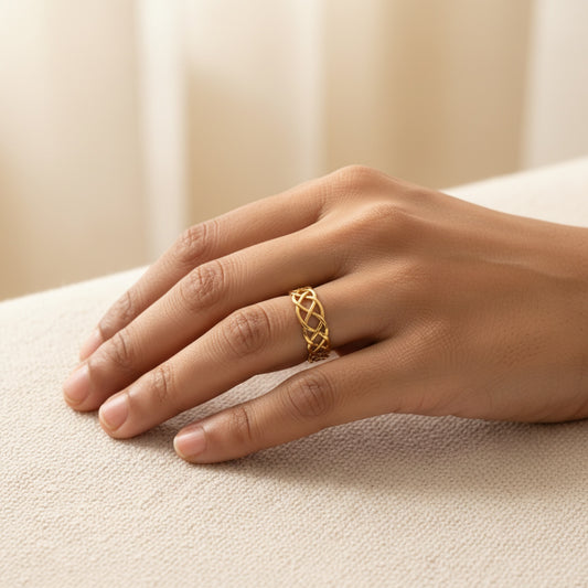A close-up of a hand wearing a delicate gold ring with an openwork, interlaced geometric pattern, resting on a textured beige fabric surface.