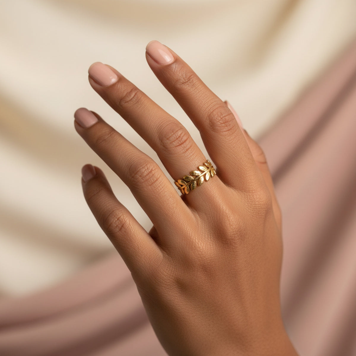 A gold laurel leaf ring worn on a model's finger against a pink satin backdrop.