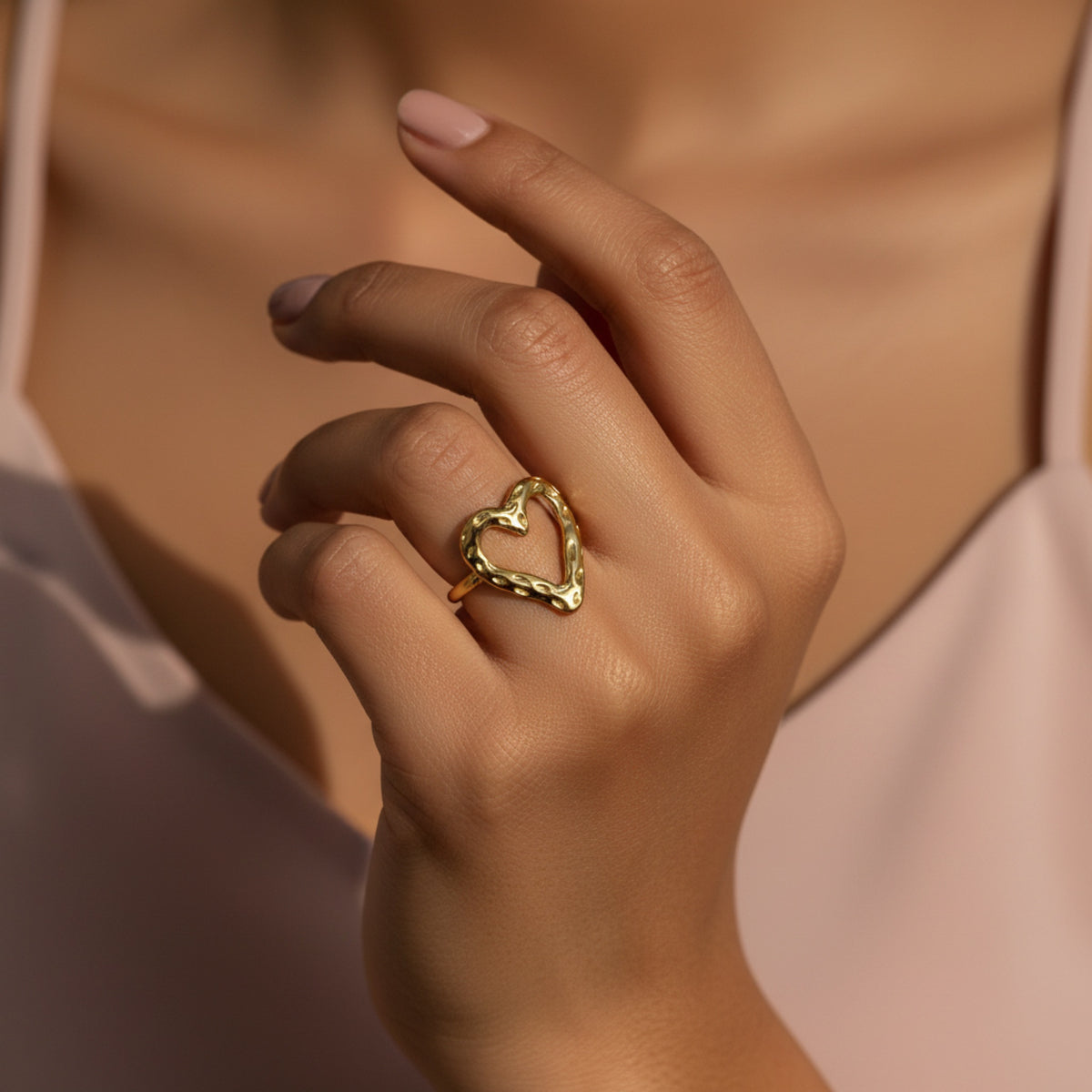 A close-up of a person's hand wearing a textured gold heart-shaped ring, set against a soft pink top and warm skin tones.