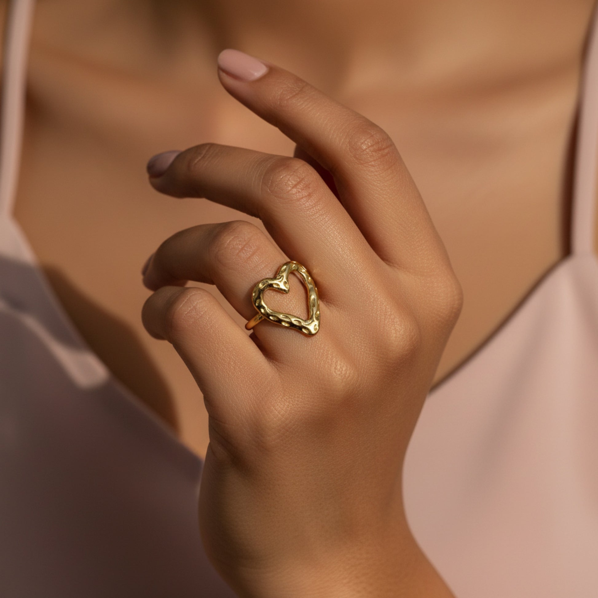 A close-up of a person's hand wearing a textured gold heart-shaped ring, set against a soft pink top and warm skin tones.
