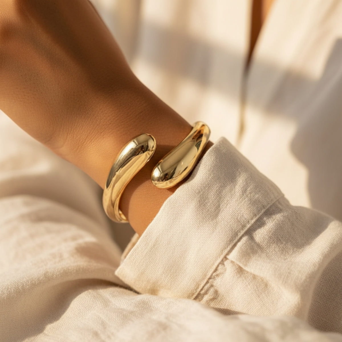 A close-up of a person's wrist wearing a polished gold split cuff bracelet, paired with a light beige linen shirt under warm, sunlit shadows.
