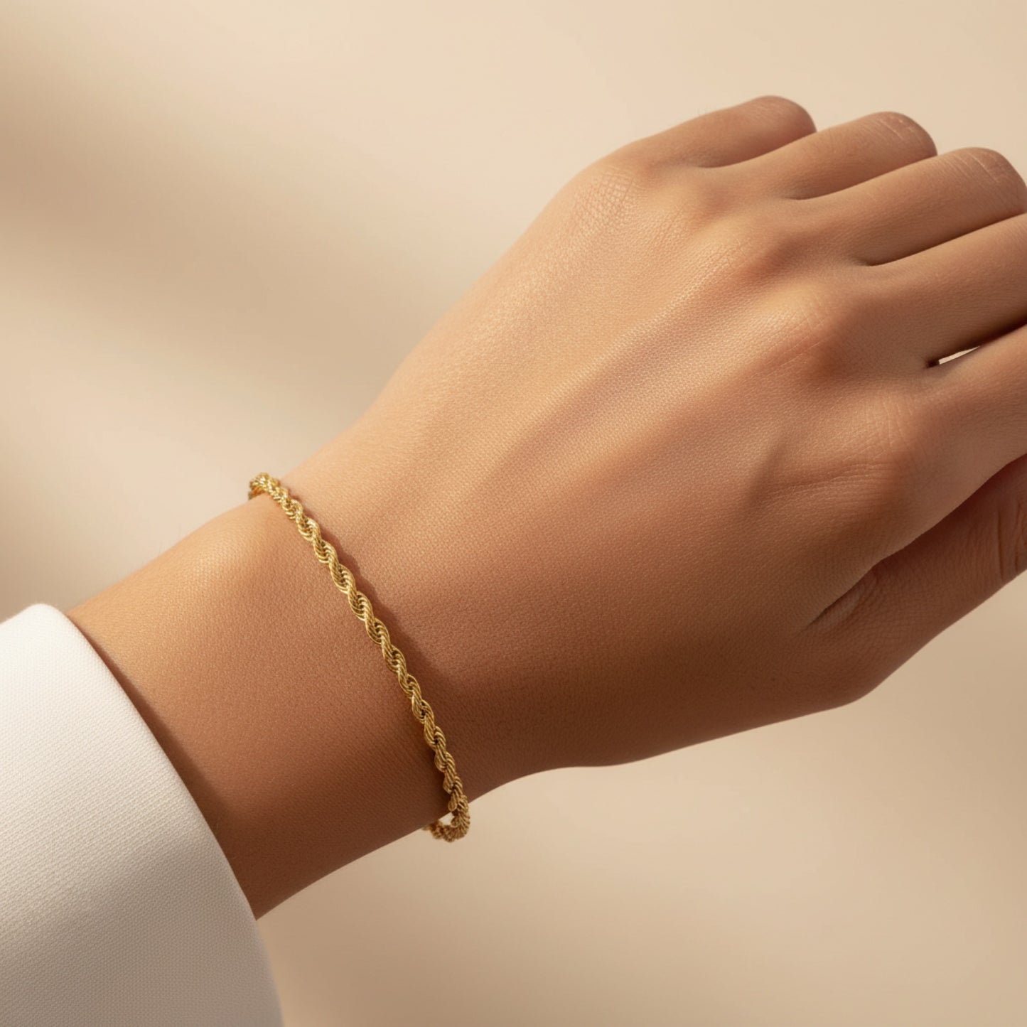 A delicate gold rope-style bracelet worn on a person's wrist, shown against a soft beige background with a white sleeve visible at the edge.