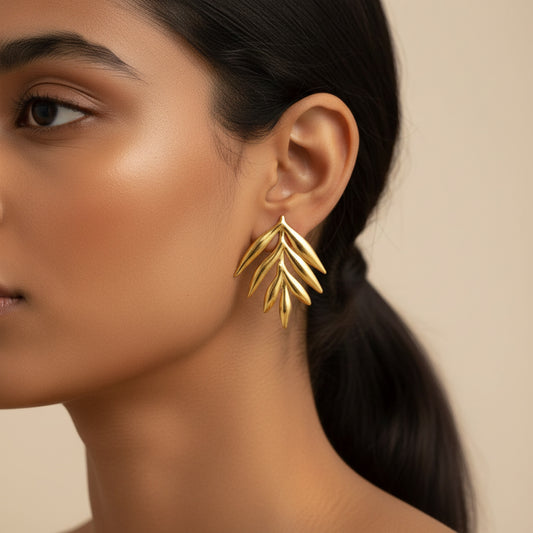 A close-up of a woman wearing a gold leaf-shaped dangle earring, showing the earring against her ear and neck, with a solid beige background.