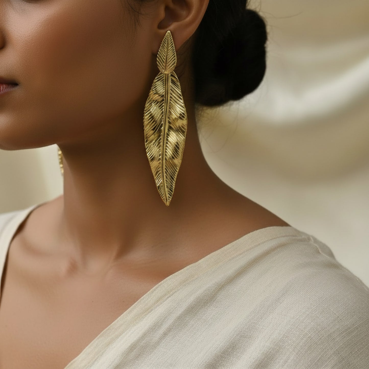 A close-up of a model wearing a long gold feather-shaped drop earring, styled with a white top against a soft beige background.