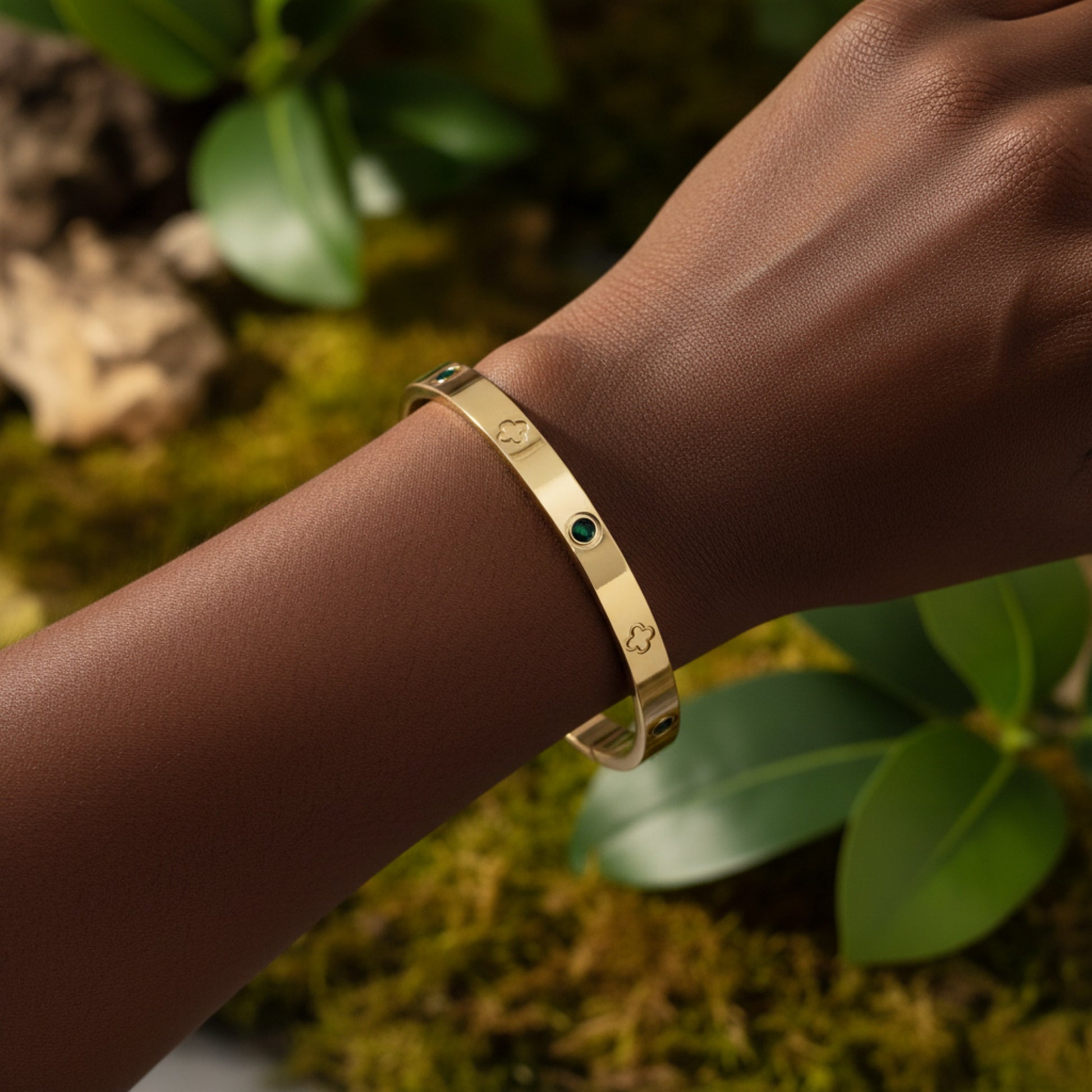 A gold bangle bracelet with small green stones and engraved clover motifs, shown worn on a person's wrist against a blurred background of green leaves and moss.