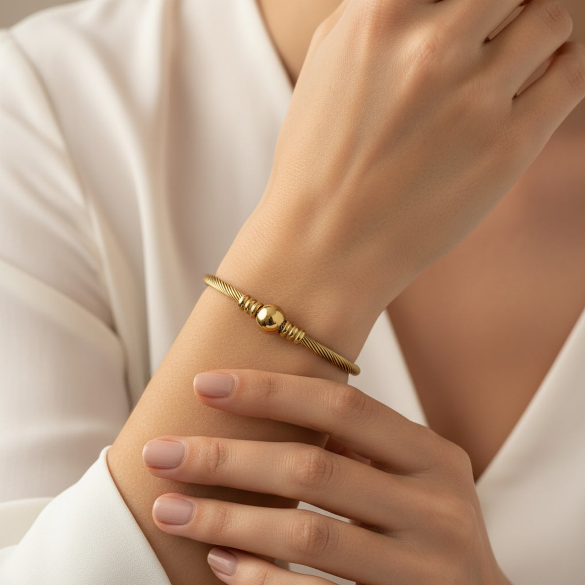 A delicate gold coiled bracelet with a central polished bead, worn on a woman's wrist. The image features a close-up of the wrist and hands, with the model wearing a white satin blouse against a soft, warm background.