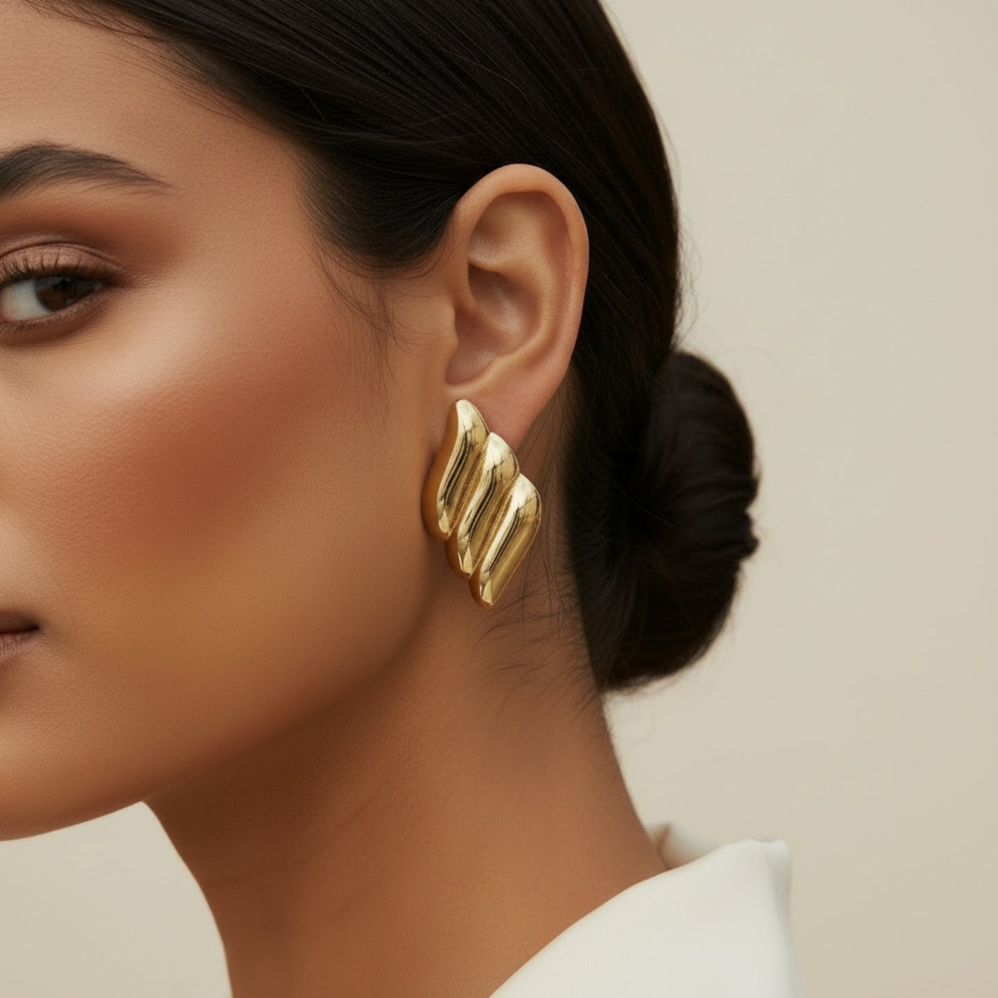 A close-up of a woman wearing a chunky, sculptural gold earring with three parallel, wavy ribbed bars, set against a soft beige background.
