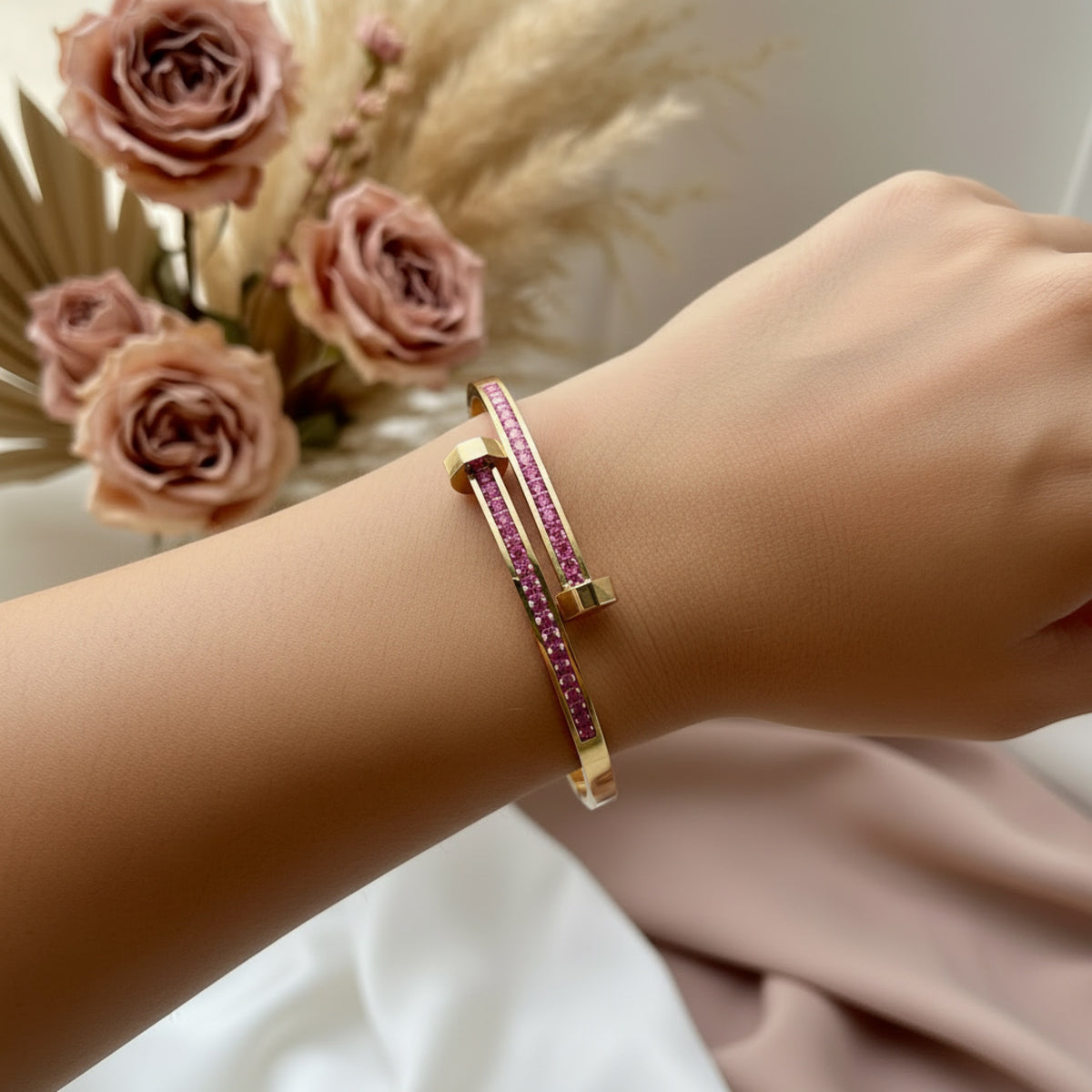 A gold-toned open cuff bracelet with a central row of pink gemstones, worn on a person's wrist against a soft neutral background with dried flowers.