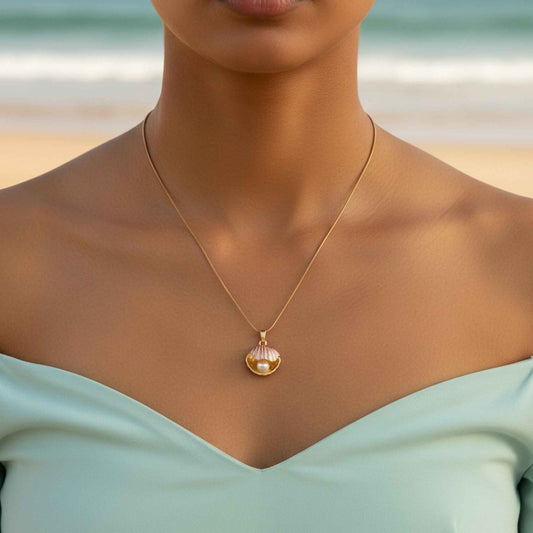 A gold pink enamel scallop shell pendant with a faux pearl inside, worn on a fine gold chain by a model in a mint dress at the beach.