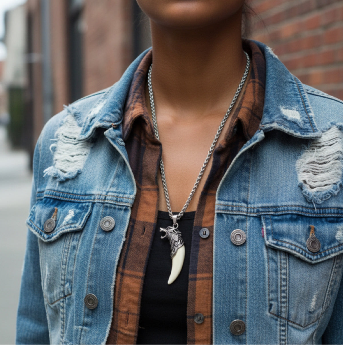 A model in a denim jacket and plaid shirt wearing a silver chain necklace with a wolf-head pendant and cream fang charm.
