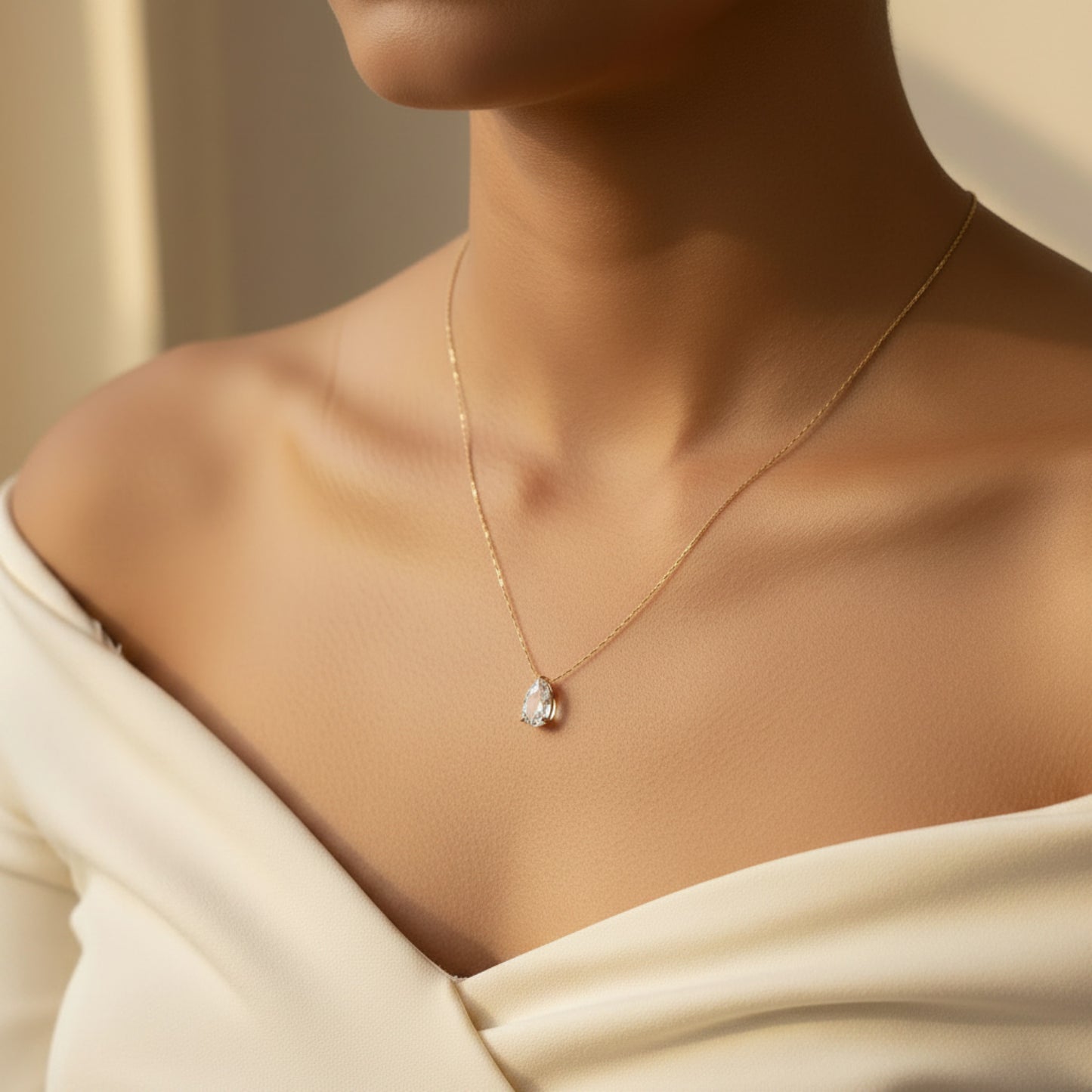 A close-up lifestyle shot of a model wearing a delicate gold chain necklace with a pear-cut clear gemstone pendant, styled with a cream off-the-shoulder top.