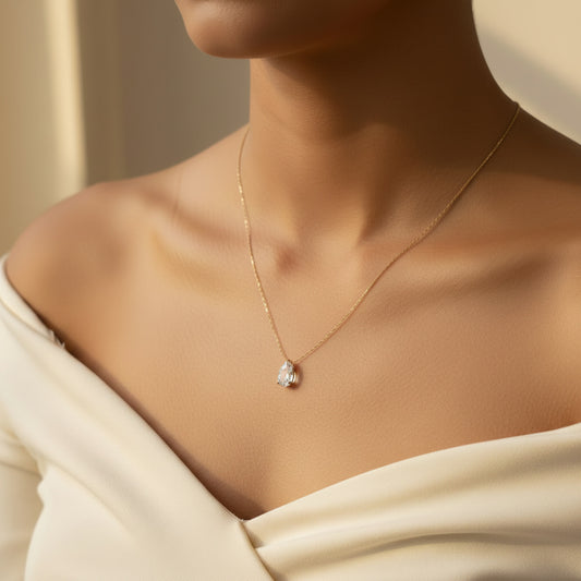 A close-up lifestyle shot of a model wearing a delicate gold chain necklace with a pear-cut clear gemstone pendant, styled with a cream off-the-shoulder top.